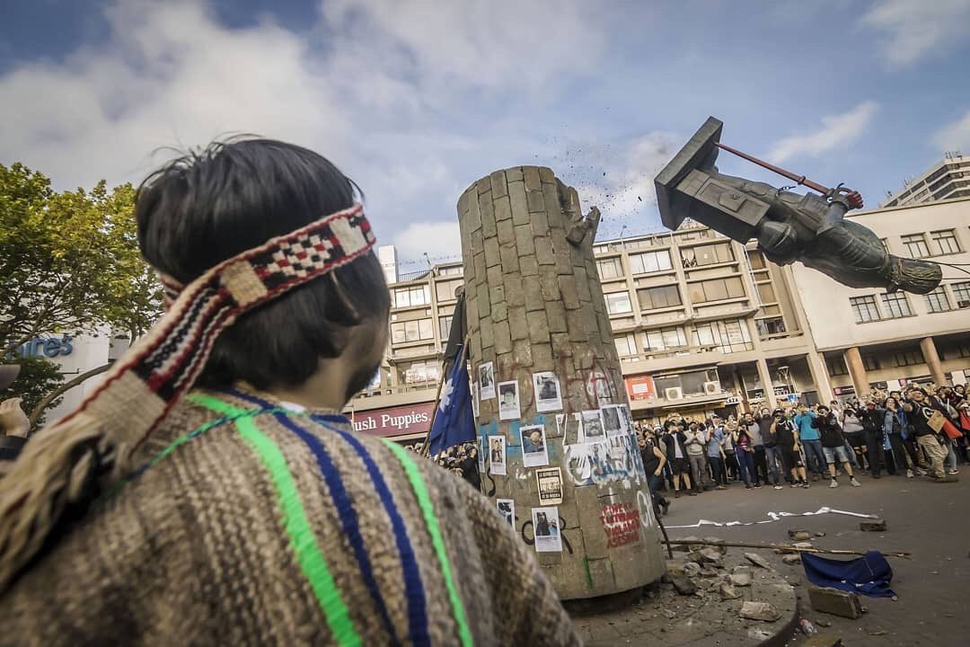 en la foto se ve una persona con vestimenta mapuche observando el monumento de un conquistador que cae, arrastrado por la fuerza de manifestantes en el contexto de la revuelta en chile en octubre 2019